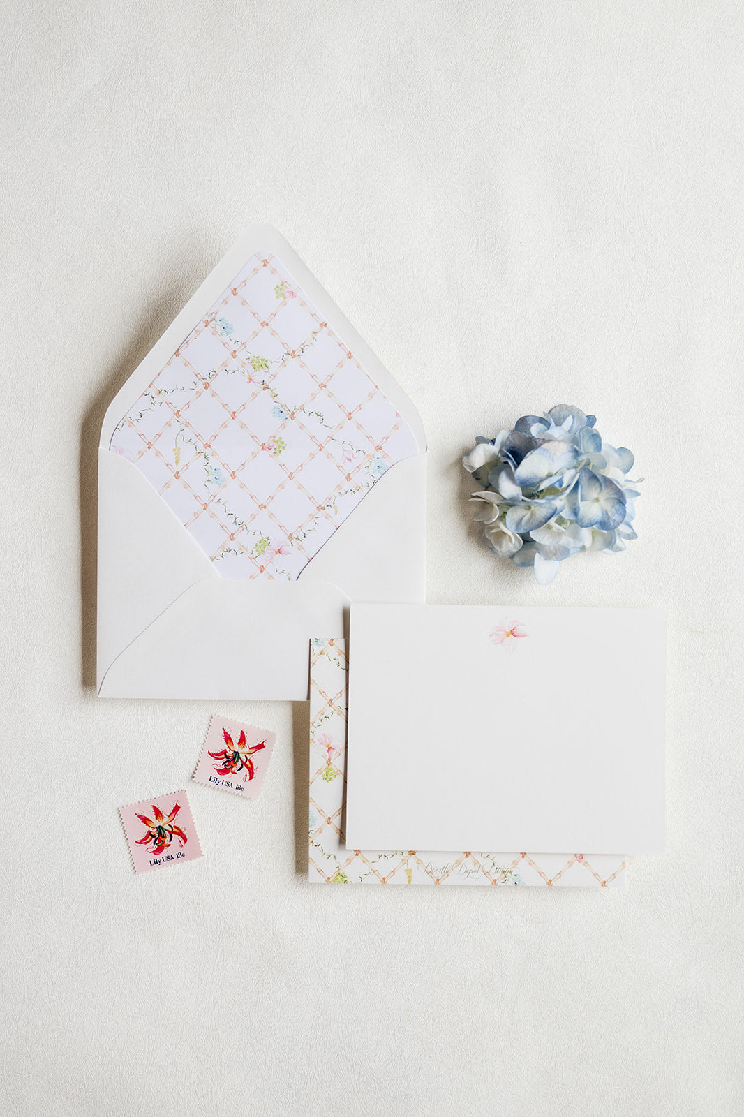 A flat-lay of a stationery suite on a white background, featuring a white envelope with a patterned interior, a blank white card, a patterned paper strip, two small floral stickers, and a blue paper flower accent.