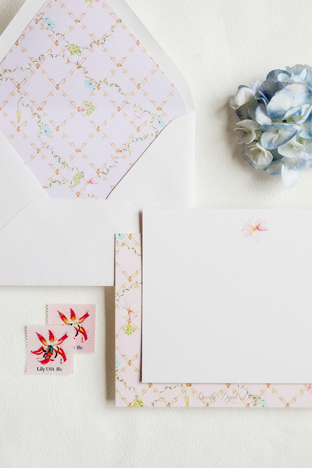 A styled flat-lay of a floral stationery set on a textured white surface, featuring a white envelope with a pale pink patterned liner, a matching patterned card, a blank white note card, two small red-and-white flower stamps, and a blue hydrangea flower.