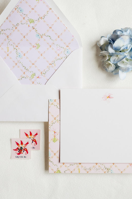 A styled flat-lay of a floral stationery set on a textured white surface, featuring a white envelope with a pale pink patterned liner, a matching patterned card, a blank white note card, two small red-and-white flower stamps, and a blue hydrangea flower.