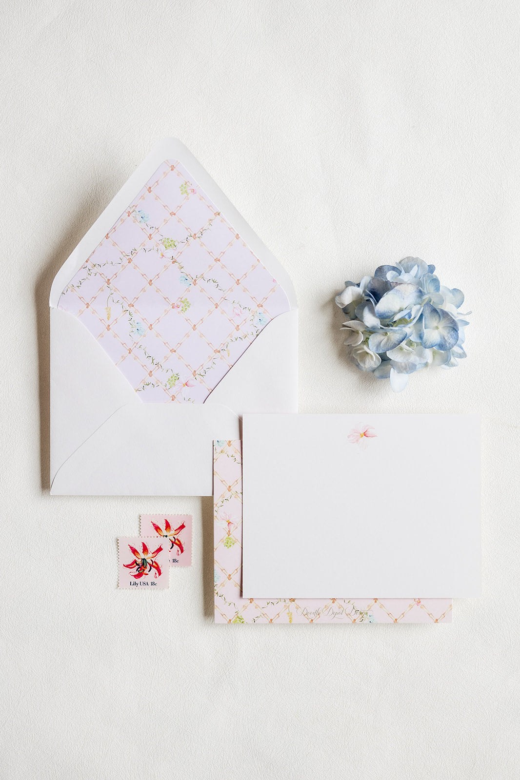 A flat-lay of stationery on a white background, featuring a white envelope with a pastel floral patterned liner, a blank white card, a matching patterned card or paper strip, two small floral stickers, and a blue hydrangea flower.