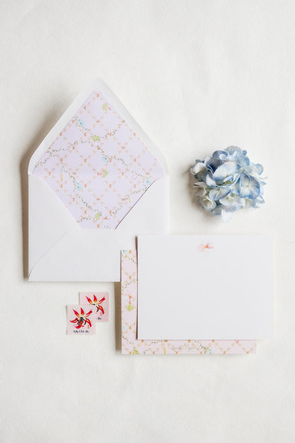 A flat-lay of stationery on a white background, featuring a white envelope with a pastel floral patterned liner, a blank white card, a matching patterned card or paper strip, two small floral stickers, and a blue hydrangea flower.